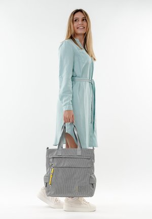 Naine helesinises vöödiga kleidis ja valgetes tossudes, hoides suurt halli käekotti kollase luku tõmbega, vaadates küljele ja naeratades.