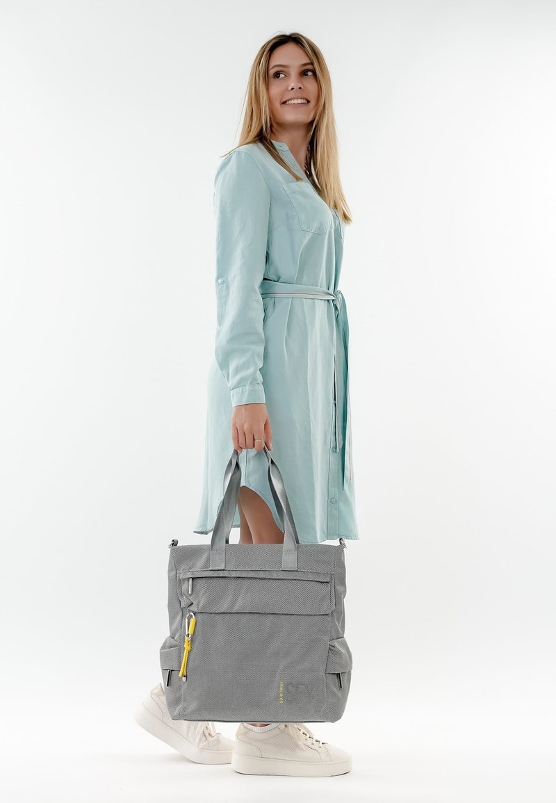 Frau in einem hellblauen, mit Gürtel versehenen Kleid und weißen Turnschuhen, die eine große graue Tragetasche mit gelbem Reißverschlussanhänger hält, seitlich blickt und lächelt.