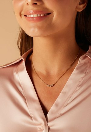 Collier en chaîne dorée avec cinq petites pierres rondes et claires disposées en ligne courbée, porté sur un chemisier en satin rose pâle.