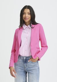 Rosa Blazer mit Frontknöpfen, gekerbtem Revers und zwei Seitentaschen, kombiniert mit einem hell gestreiften Hemd und abgenutzten blauen Jeans.