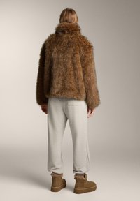 Barna műszőrme dzseki magas gallérral, világosszürke melegítőnadrággal és barna bokacsizmával párosítva. A dzseki puhanak és texturáltnak tűnik.