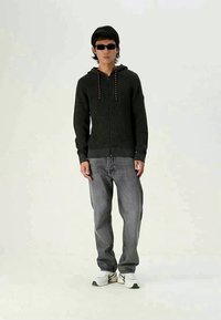 Σκούρο πράσινο πλεκτό φούτερ με κόκκινες κορδόνες, συνδυασμένο με γκρι φαρδιά τζιν και λευκά αθλητικά παπούτσια με πράσινες λεπτομέρειες. Το μοντέλο φοράει γυαλιά ηλίου.