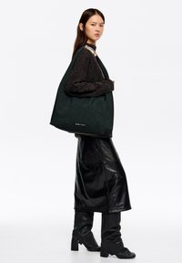 Duża, ciemnozielona, pikowana torba na ramię z fakturowanej tkaniny z paskiem na ramię. Modelka ubrana w czarny, oversize'owy sweter oraz błyszczącą czarną spódnicę.