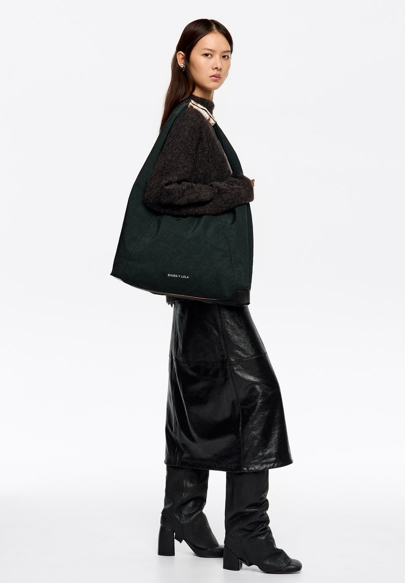 Duża, ciemnozielona, pikowana torba na ramię z fakturowanej tkaniny z paskiem na ramię. Modelka ubrana w czarny, oversize'owy sweter oraz błyszczącą czarną spódnicę.