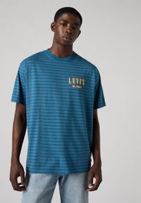 T-shirt azul às riscas feita de algodão, com decote redondo e um logo amarelo "LEVI'S" no lado esquerdo do peito. Modelo a usar um jeans claro.
