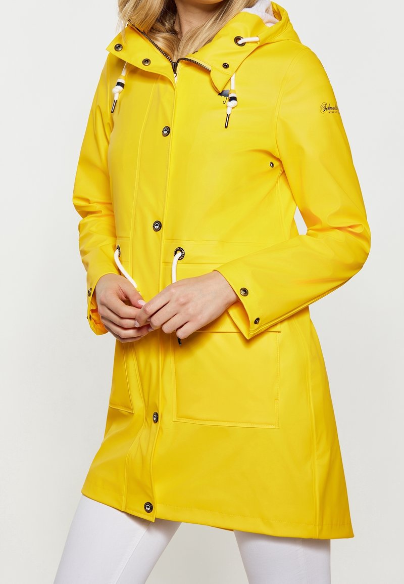 Femme portant un imperméable jaune vif avec capuche, boutons sur le devant, taille resserrée par un cordon et grandes poches avant.