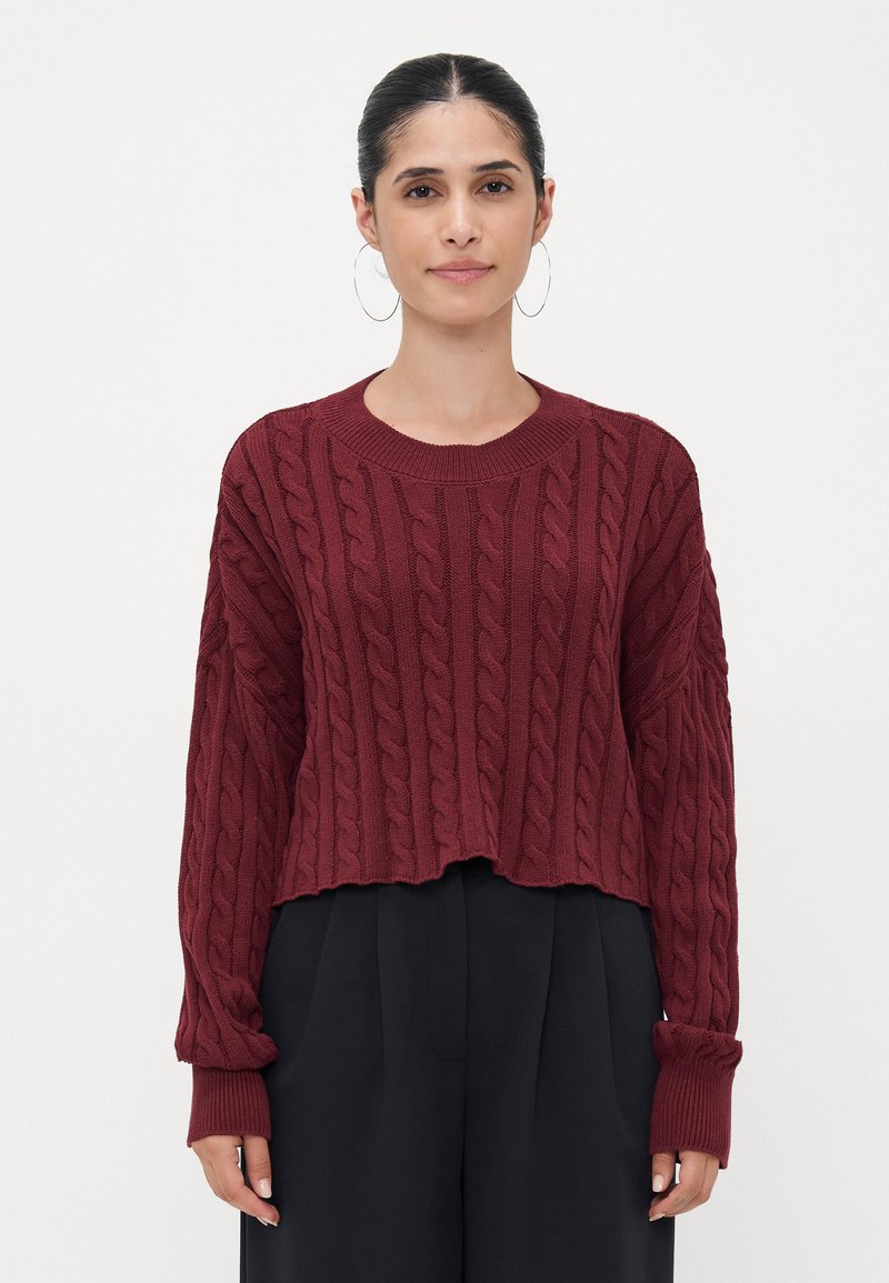 Burgundy cable-knit sweater with a cropped design, wide sleeves, and ribbed cuffs, paired with black, loose-fitting trousers.