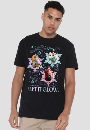 Young man wearing a black T-shirt featuring three animated female characters with floral and musical designs and text "HUNTRX LET IT GLOW."
