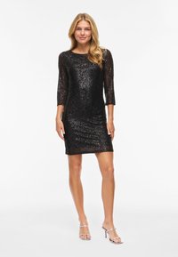 Blondynka stoi w czarnej, cekinowej sukience do kolan z rękawami do łokci i srebrnych sandałach na wysokim obcasie.