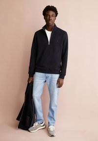 Schwarzer Zip-Hoodie mit hohem Kragen, kombiniert mit hellblauen Jeans und grauen Sneakers, gehalten mit einer schwarzen Jacke. Schlichtes, lässiges Design.