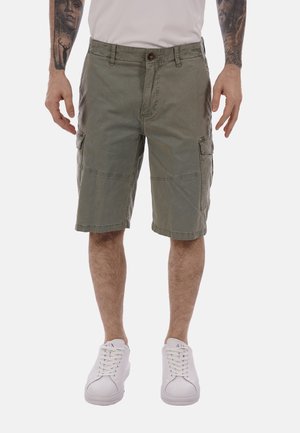 Uomo che indossa pantaloncini cargo verde oliva con multiple tasche, scarpe da ginnastica bianche e una maglietta bianca, mostrando le braccia tatuate.