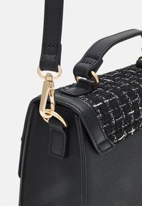 Musta käsilaukku, jossa teksturoitu mustavalkoinen kudottu yläosa, jämäkkä muoto, kultainen lukko ja säädettävä nahkahihna.