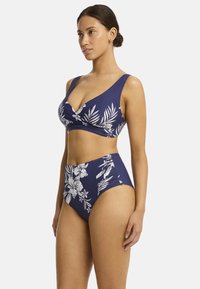 Kobieta pozująca w granatowym bikini w kwiatowy wzór z wysokim stanem, prezentując pewny siebie wyraz twarzy i zrelaksowaną postawę.