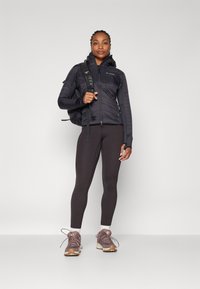 Giacca nera isolata con cappuccio, design aderente, abbinata a leggins scuri e scarpe da trekking texturizzate. Zaino con spallacci regolabili.