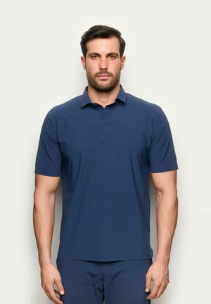 Mann mit kurzen dunklen Haaren und Bart, der ein schlichtes marineblaues Polohemd mit kurzen Ärmeln trägt und vor hellem Hintergrund steht.
