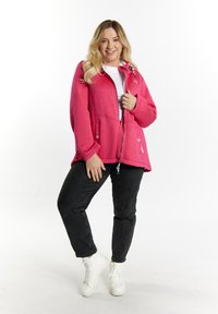 Giacca in pile di un vivace colore rosa con chiusura a zip, tasche laterali e dettagli decorativi; abbinata a jeans neri e sneakers bianche.