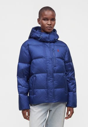Person iført en blå vatteret dunjakke med hætte og lynlåslomme på brystet, kombineret med lyseblå jeans, stående mod hvid baggrund.