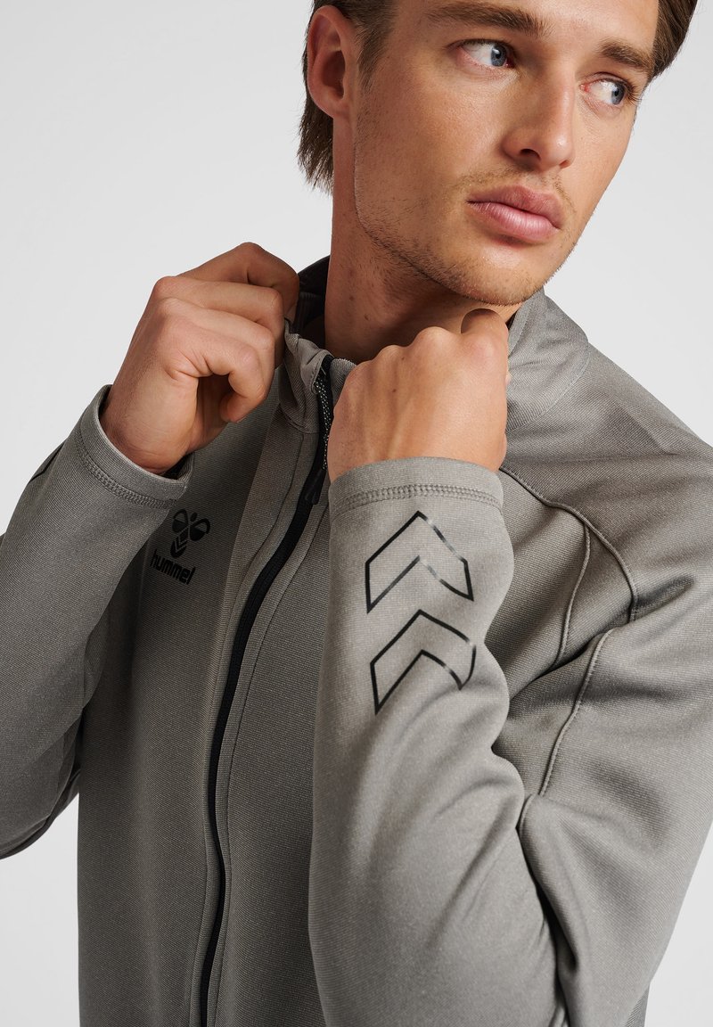 Grau-braune Zip-Jacke aus strukturiertem Material, mit schwarzen Chevron-Designs an den Ärmeln und einem Logo auf der Brust. Nahaufnahme des Kragens.