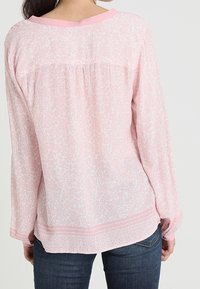 Blusa rosa chiaro a maniche lunghe con motivo floreale, scollatura rotonda e colletto in contrasto. Orlo leggermente curvo e dettaglio arricciato sul retro.