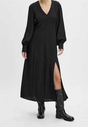 Schwarzes Maxikleid mit V-Ausschnitt, langen Puffärmeln und einem hohen Schlitz bis zum Oberschenkel. Hergestellt aus glattem Stoff, der einen dezenten Glanz hat. Getragen mit schwarzen kniehohen Stiefeln.