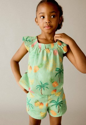 Jeune fille vêtue d'un haut sans manches vert et d'un short avec des motifs colorés de fruits tropicaux et de palmiers, regardant sur le côté avec une main sur la hanche.