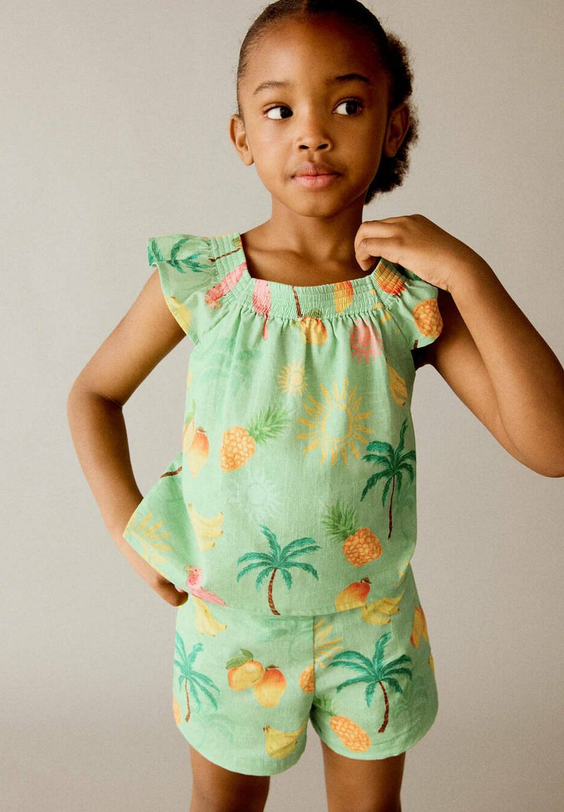 Jeune fille vêtue d'un haut sans manches vert et d'un short avec des motifs colorés de fruits tropicaux et de palmiers, regardant sur le côté avec une main sur la hanche.