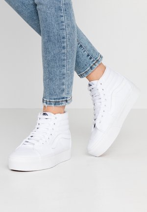 Jambes en jean denim bleu portant des baskets montantes blanches à semelles épaisses sur un sol et un fond gris clair.