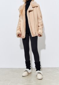 Giacca beige foderata in pelliccia sintetica con ampio colletto, abbinata a leggings neri e scarpe slip-on color crema con dettagli in pelliccia e hardware dorato.