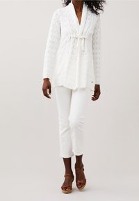 Cardigan bianco in pizzo con colletto a scialle, vita legata e maniche lunghe, abbinato a pantaloni bianchi e sandali marroni con lacci.