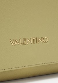 Superficie in pelle testurizzata verde chiaro con lettering dorato "VALENTINO" e cuciture visibili vicino al margine inferiore.