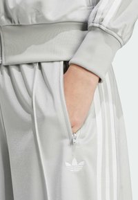 Vale harmaa urheilupuku, jossa on ribattu hihansuu, varustettuna vetoketjupussilla ja kolmella valkoisella raidalla sivuilla. Adidas-logo pussissa.