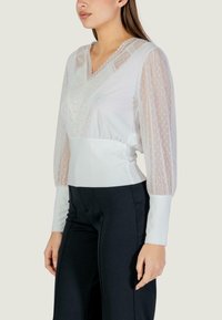 Biała bluzka z przezroczystymi rękawami w groszki, dekoltem w kształcie litery V i misternym zdobieniem z koronki. W połączeniu z czarnymi spodniami. Gładka tkanina i dopasowany krój.