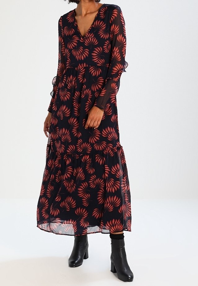 Femme portant une longue robe noire avec des motifs de feuilles rouges, des manches longues transparentes et des bottines noires, debout devant un fond uni.