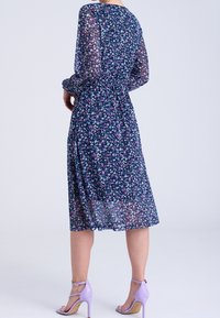 Kukallinen mekko tummansinisellä taustalla, jossa on pitkät, läpikuultavat hihat, tiukka vyötärö ja virtaava helma. Mekossa on pieniä monivärisiä kukkakuosisia kuvioita.