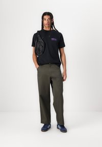 Zwart katoenen T-shirt met paarse graphic, olijfgroene wijde broek, en blauwe sneakers met lichtblauwe strepen, uitgerust met een zwarte schoudertas.