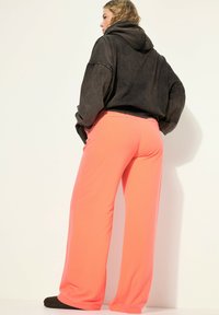 Černá oversized mikina s kapucí a širokými rukávy, spárovaná s vysokými, splývavými kalhotami neonově růžové barvy, které mají hladkou texturu a žádné viditelné vzory.