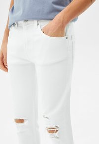 Jeans de denim blanco con un diseño ajustado, que presentan detalles desgastados en las rodillas. La tela tiene una textura suave y un acabado brillante.