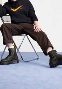 Svarta stövlar med grön sömnad och gula detaljer, med tjock sula och högt skaft, kombinerade med mörkbruna byxor och en svart huvtröja.