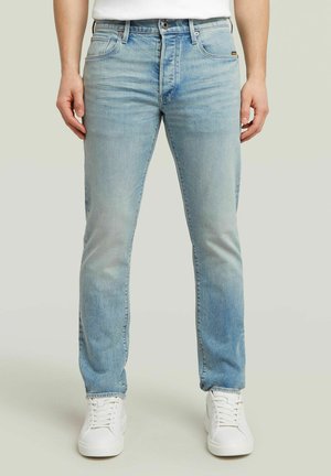 Homme portant un jean slim bleu clair et des baskets blanches, se tenant devant un fond neutre.