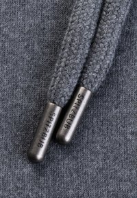 Graues Strickgewebe mit zwei kontrastierenden grauen Kordeln. Die Kordeln haben Metallenden, die mit "SPITZBUB" beschriftet sind. Die Textur ist glatt und gewebt.