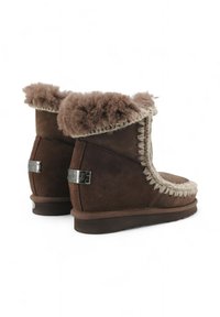 Stivali ankle in suede marrone con una morbida fodera in pelliccia tan, dettagli intrecciati lungo il bordo e un piccolo accento di logo metallico sul lato.