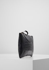 Sac à main en cuir noir avec un motif crocodile texturé, un rabat supérieur arrondi et une anse unique. Forme rectangulaire avec des accents lisses.