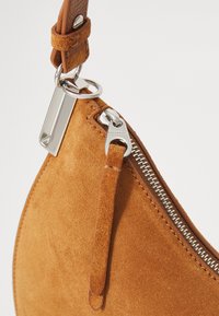 Borsa a spalla in suede beige con zip argentata, tracolla in pelle e gancio in metallo. Presenta una forma curva e una texture morbida.