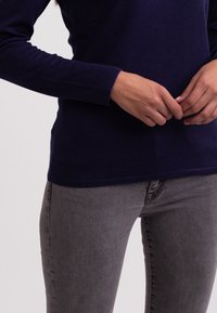 Granatowa bluzka z długim rękawem, wykonana z miękkiego materiału, połączona z dopasowanymi szarymi dżinsami o delikatnej fakturze i standardowych detalach kieszeni.