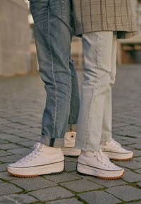 Vita hög-top sneakers med texturerade gummisulor, svarta ränder och grå detaljer, som bärs med ljusa och mörka tvättade jeans på en kullerstensyta.