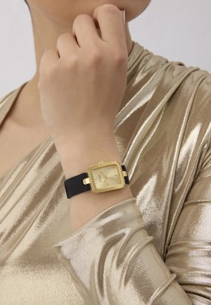 Personne portant un haut en tissu doré brillant avec une montre carrée dorée avec un bracelet noir au poignet gauche, main près du menton.