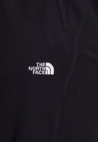 Schwarzer Fleece-Stoff mit Struktur, versehen mit einem gestickten weißen Logo von "The North Face" auf der linken Seite. Keine weiteren Details vorhanden.