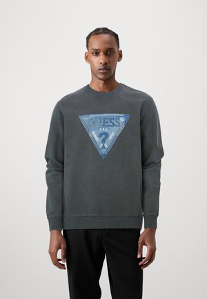 Sweatshirt gris avec une appliqué triangulaire en denim bleu portant le logo "GUESS" et le texte "U.S.A.". Bords côtes aux poignets et à l'ourlet.