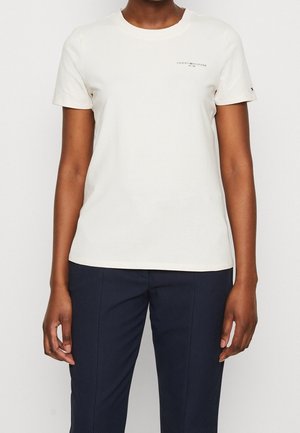 Person iført en ensfarvet hvid T-shirt med korte ærmer og lille logo på brystet, kombineret med mørke teksturerede bukser, stående mod en hvid baggrund.