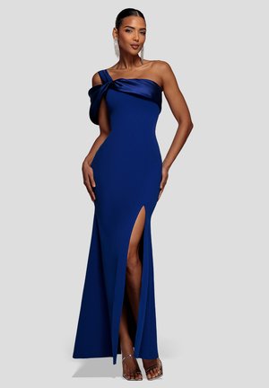 Mujer con un vestido azul marino largo hasta el suelo, de un solo hombro, con una abertura en el muslo y sandalias plateadas de tacón alto, posando frente a un fondo gris claro.
